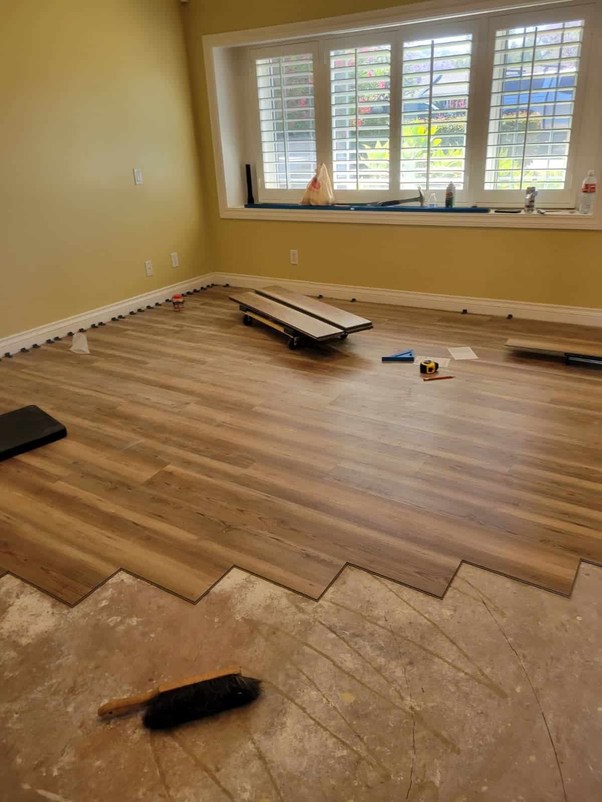 Newly installed hardwood flooring in living room with ongoing maintenance, tools, and construction materials visible, bright window with shutters in the background.
