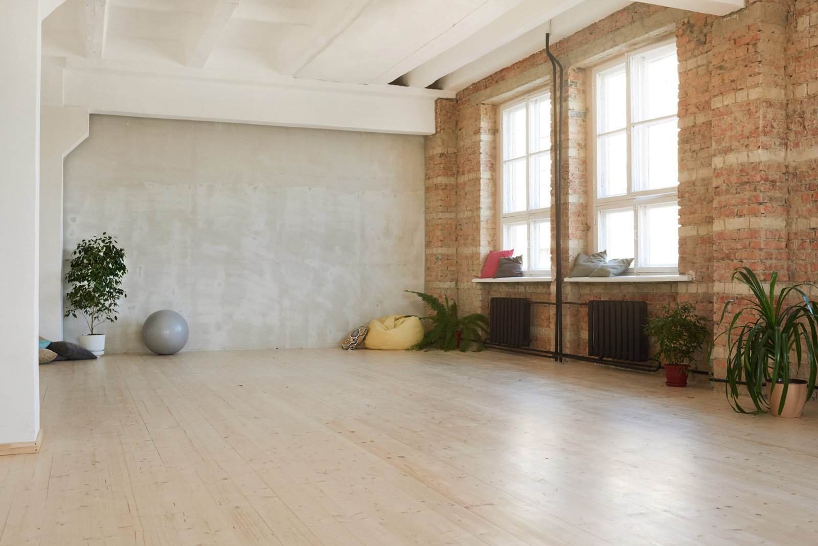 Modern industrial-style living space with exposed brick walls, large windows, and light wooden flooring, featuring minimal decor and indoor plants.